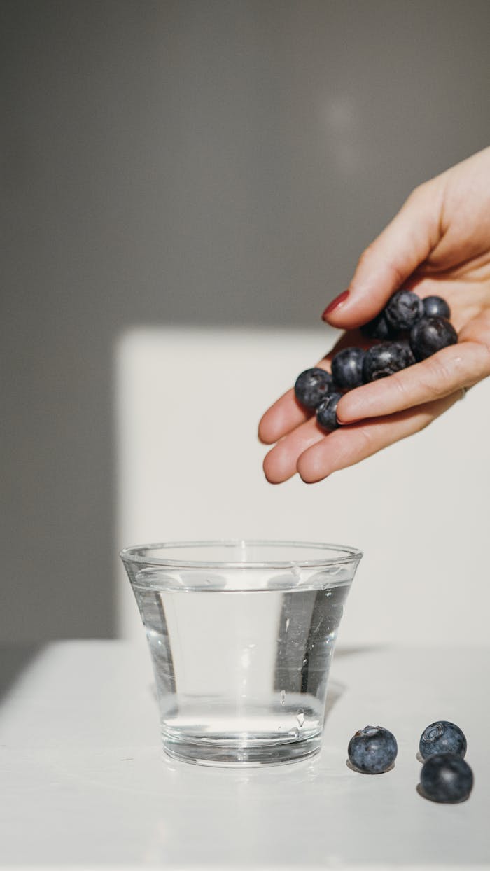 creative-02 A hand holding ripe blueberries above a glass of water, emphasizing freshness and health.